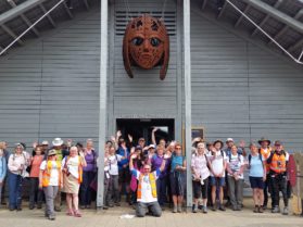 A large group of walkers cheering outside the exhibit hall at Sutton Hoo with Raedwald's rust-orange mask looking down on them