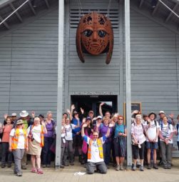 A large group of walkers cheering outside the exhibit hall at Sutton Hoo with Raedwald's rust-orange mask looking down on them