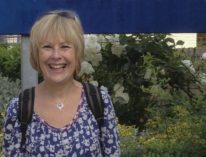Image of Leslie Dolphin in a blue and white floral top. She is smiling and standing in front of a large white rose bush and a blue sign